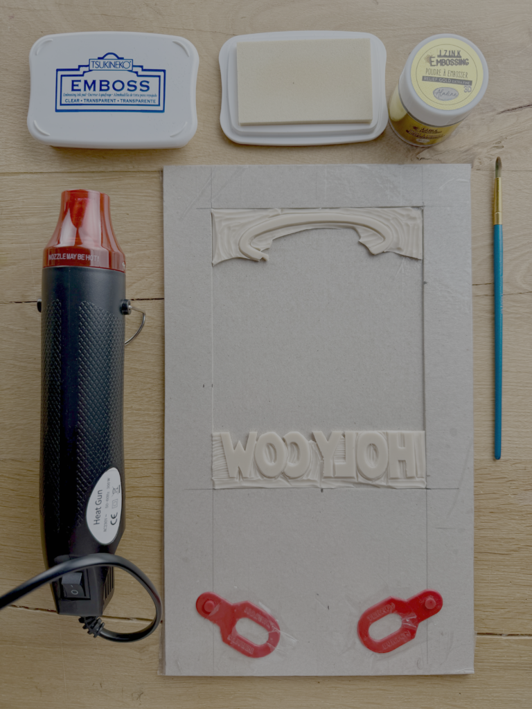 Materials used for adding gold embossing to a linoprint, including clear embossing ink, gold embossing powder and a mini heat gun.
